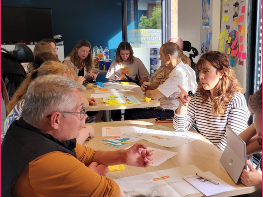Deux tables au centre d'une dizaine de personnes qui discutent et réflechissent. Derrière, un mur de post-it