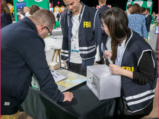 Deux personnes à un stand découvrent sur une table un live game composé d'une boîte et d'une carte, présentée par une troisième personne